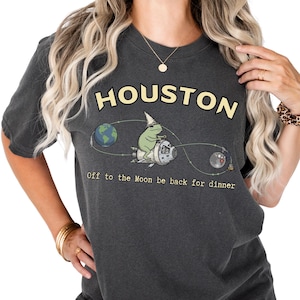 May include: Dark gray t-shirt with the word "HOUSTON" in yellow block letters. A cartoon frog in a wizard hat rides a rocket ship toward the moon. The text "Off to the Moon be back for dinner" is below the graphic.
