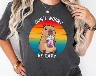 May include: Dark gray t-shirt featuring a cartoon capybara drinking a pink bubble tea. The design is set against a colorful sunset background with the text "DON'T WORRY BE CAPY" in white. A casual, comfortable tee.