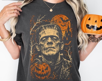Könnte beinhalten: Ein anthrazitfarbenes T-Shirt mit einem Grafikdruck von Frankensteins Monster, einem Kürbis, einem Vollmond, Fledermäusen und der Silhouette eines Spukhauses. Das Design ist in Orange- und Gelbtönen gehalten. Das Wort Pepper ist in einem Kasten gedruckt.