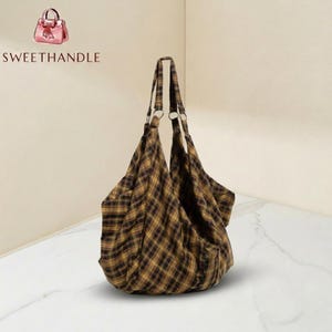 Könnte beinhalten: Eine große, braun-gelb karierte Tragetasche mit zwei Griffen und silbernen Metallringen. Die Tasche hat ein breites, offenes Design und ist aus einem weichen Stoff gefertigt. Der Markenname "SWEETHANDLE" ist oben links sichtbar.