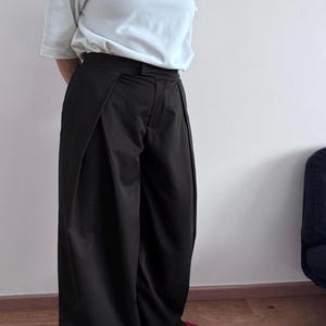 May include: Black wide-leg pants with a high waist and a white t-shirt. The pants have a unique design with pleats at the front. The person is wearing red shoes. The outfit is simple and stylish.
