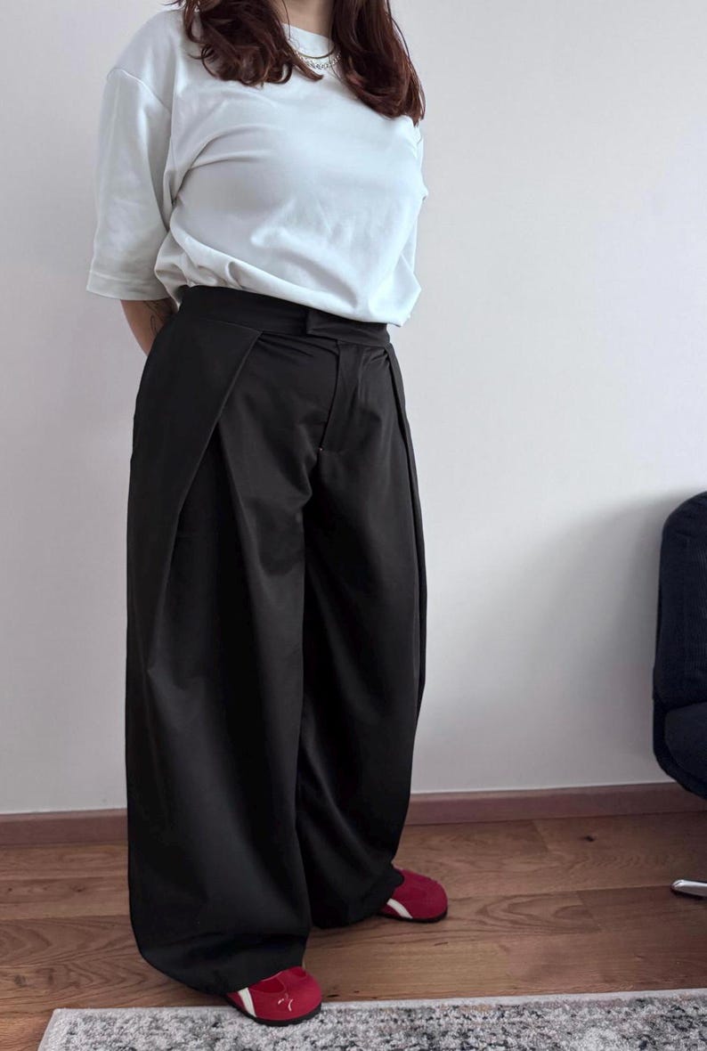 May include: Black wide-leg pants with a high waist and a white t-shirt. The pants have a unique design with pleats at the front. The person is wearing red shoes. The outfit is simple and stylish.