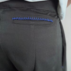 May include: Close-up of black pants with a blue decorative stitch detail on the back pocket. The pants are made of a smooth fabric and appear to be tailored. The stitching adds a pop of color.