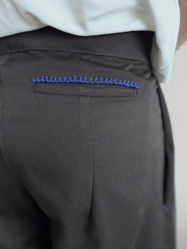May include: Close-up of black pants with a blue decorative stitch detail on the back pocket. The pants are made of a smooth fabric and appear to be tailored. The stitching adds a pop of color.