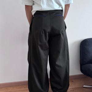 May include: Dark olive green wide-leg pants with a high waist and a relaxed fit. The pants have large pockets and a blue accent on the waistband. The person is wearing a white t-shirt and black shoes.