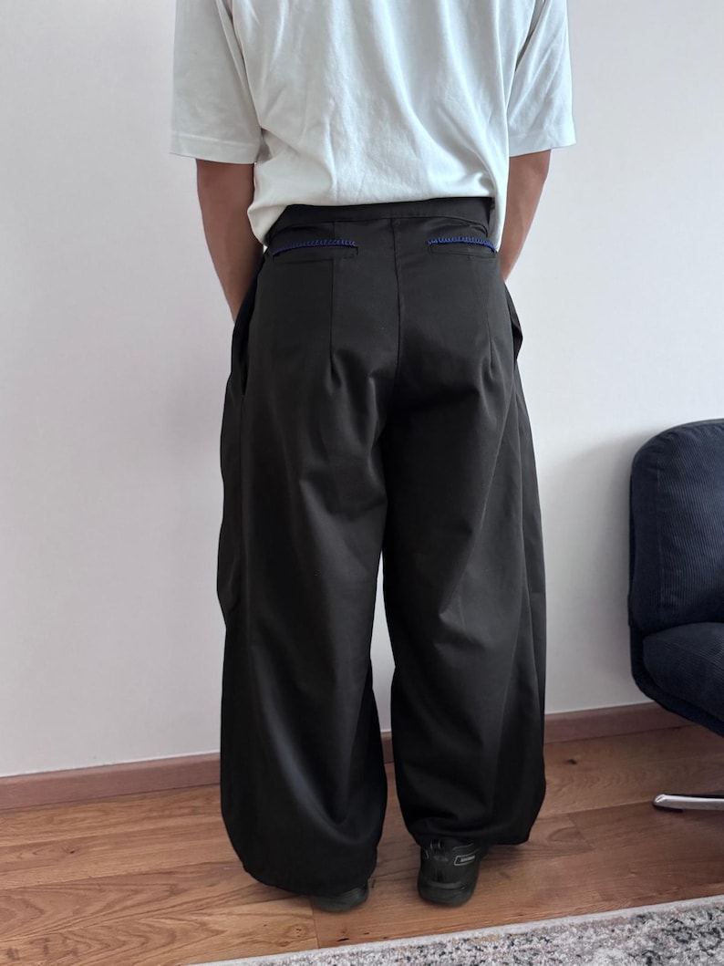 May include: Dark olive green wide-leg pants with a high waist and a relaxed fit. The pants have large pockets and a blue accent on the waistband. The person is wearing a white t-shirt and black shoes.