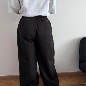 May include: Black wide-leg pants with red stitching detail at the back pockets. The pants are paired with a white t-shirt. The outfit is displayed from the back.