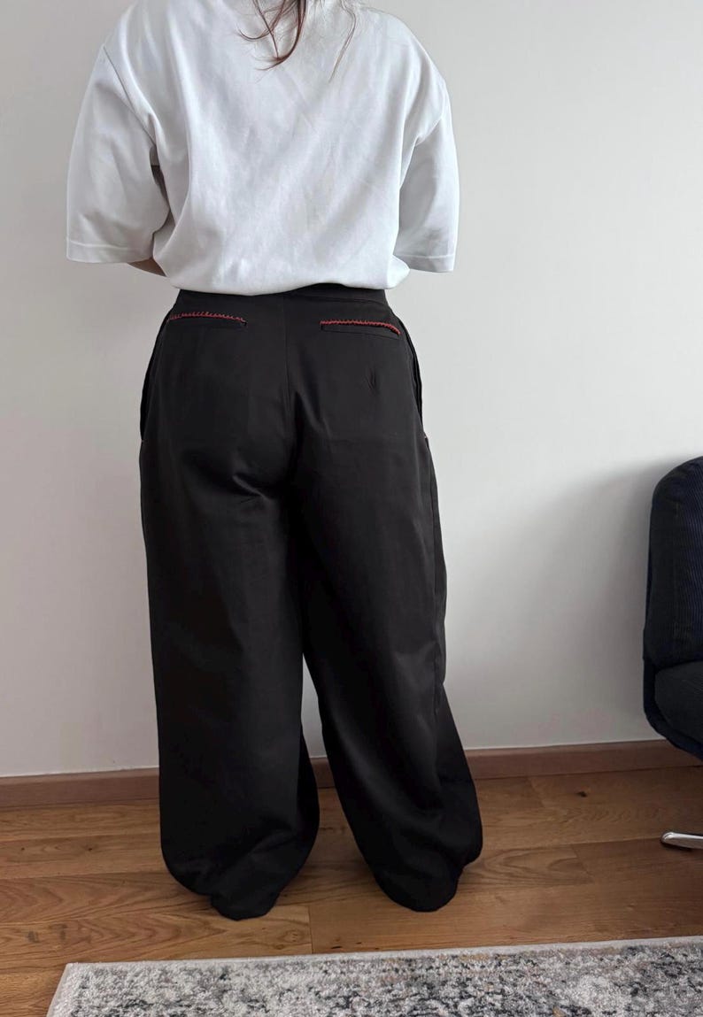 May include: Black wide-leg pants with red stitching detail at the back pockets. The pants are paired with a white t-shirt. The outfit is displayed from the back.