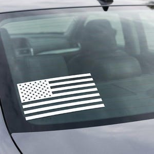 May include: A white vinyl decal of the United States flag applied to the rear window of a dark gray sedan. The decal features the iconic stars and stripes design in a solid white color, contrasting against the dark tinted glass of the vehicle.