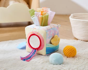 May include: A colorful baby activity cube with various textures and features. The cube has a mirror, a starfish, and a sea-themed design. It includes soft, colorful tissue-like pieces and two small balls. The cube is designed for sensory play.