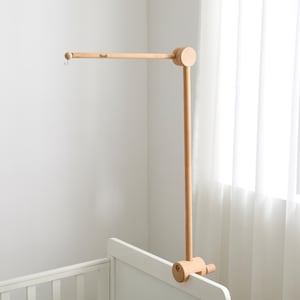 Könnte beinhalten: Ein Baby-Mobile-Arm aus Naturholz, befestigt an einem weißen Kinderbett. Der Mobile-Arm hat eine horizontale Stange mit einem kleinen Haken und eine vertikale Stütze. Das Design ist minimalistisch und modern.