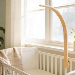 Peut inclure: Un bras de mobile pour bébé en bois clair fixé à un lit bébé blanc. Le bras incurvé s'étend au-dessus du lit, prêt à accueillir un mobile. Le lit est près d'une fenêtre avec des rideaux transparents. Une couverture beige est drapée sur le lit.