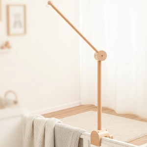 May include: A wooden baby mobile arm in a natural finish. The arm is attached to a clamp that secures to a crib. The mobile arm has a long, angled support with a small hook at the end for hanging baby toys. The design is simple and minimalist.