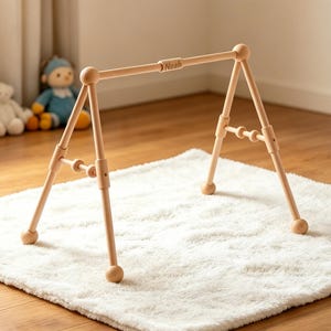 May include: A natural wood baby play gym with the name "Noah" engraved on the top bar. The gym has a triangular frame with rounded feet and is set on a white, fluffy rug. Soft toys are visible in the background.