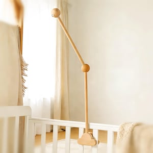 May include: A wooden baby mobile arm in a natural finish, attached to a white crib. The mobile arm has a curved design with a ball joint and a cloud-shaped clamp. The top has a hook for hanging a mobile.