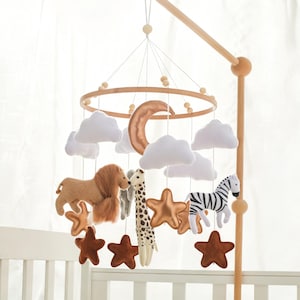 May include: A nursery mobile featuring a circular wooden frame suspended from a wooden arm. It includes plush white clouds, a metallic bronze crescent moon, bronze stars, brown stars, and stuffed animal figures of a lion, giraffe, and zebra.