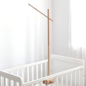 May include: A wooden baby mobile stand attached to a white crib. The stand is made of light-colored wood and features a horizontal arm extending from the top. The crib has a slatted design.