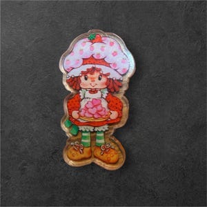 May include: A vintage-style Strawberry Shortcake character pin. The pin features a smiling girl in a red dress, white apron, and a strawberry-adorned bonnet. She holds a plate of heart-shaped treats. The pin has a gold-colored border.