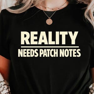 May include: Black t-shirt with the white text "REALITY NEEDS PATCH NOTES". The shirt is knotted at the waist. A patterned jacket is worn over the shirt, paired with blue jeans.
