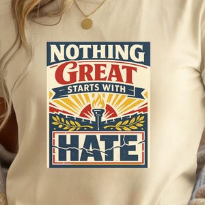 Könnte beinhalten: Ein cremefarbenes Kurzarm-T-Shirt mit einem Grafikdesign, das den Text "NOTHING GREAT STARTS WITH HATE" in Marineblau und Rot zeigt. Die Illustration enthält eine zentrale Fackel, Sonnenstrahlen und Lorbeerzweige in Marineblau, Rot und Gelb.
