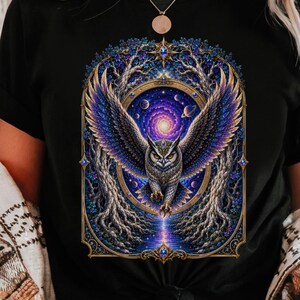Könnte beinhalten: Ein schwarzes T-Shirt mit einem Grafikdruck einer Eule mit weit ausgebreiteten Flügeln vor einem kosmischen Hintergrund mit wirbelnden violetten und blauen Nebelmustern, umrahmt von goldenen Ästen und einem Stern an der Spitze.