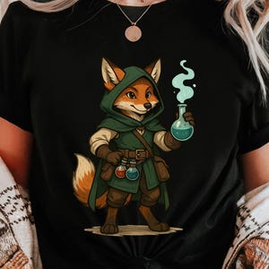 May include: Black t-shirt featuring a cartoon fox dressed as an alchemist, wearing a green hooded cloak and holding a flask with blue liquid and green vapor. The fox has orange fur and is standing upright.