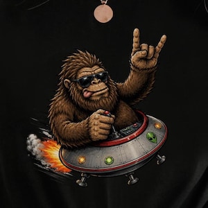 Könnte beinhalten: Ein schwarzes T-Shirt mit einer Grafik eines braunen Bigfoot, der eine schwarze Sonnenbrille trägt, die Zunge herausstreckt und eine Rock-and-Roll-Handgeste macht, während er eine silberne fliegende Untertasse steuert, die Flammen und Rauch ausstößt.