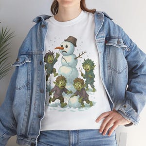 Puede incluir: Camiseta blanca con una ilustración caprichosa de un muñeco de nieve con expresión triste, con sombrero y rodeado de traviesas criaturas verdes. Las criaturas se representan en varias poses, algunas sosteniendo bolas de nieve. La obra de arte está ambientada sobre un fondo nevado.