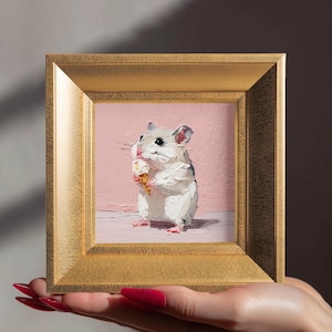 Könnte beinhalten: Gerahmtes Gemälde eines Hamsters, der ein Eis isst. Das Kunstwerk hat einen goldenen Rahmen und einen rosa Hintergrund. Der Hamster ist weiß und grau mit rosa Füßen und hält die Eistüte.