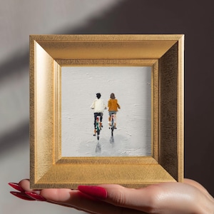 Puede incluir: Pintura enmarcada que representa a dos personas en bicicleta, alejándose del espectador. Una lleva una chaqueta blanca, la otra una chaqueta naranja. La obra está enmarcada en un marco dorado, sobre un fondo gris.