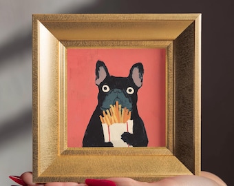 May include: A square painting in a gold frame featuring a black French bulldog with wide eyes holding a white carton of golden-brown french fries against a solid coral-pink background. The artwork is presented in a textured metallic gold frame.