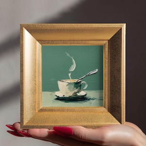 May include: A small, square painting in a gold frame depicts a cup of coffee on a saucer with a spoon. The coffee cup is white, and steam rises from the coffee. The background is a teal colour.