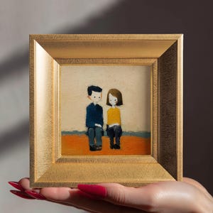 May include: A small, framed painting featuring two stylized figures seated side-by-side. The figures are depicted with simple shapes and colors: one in a blue shirt, the other in yellow. The frame is gold-colored.