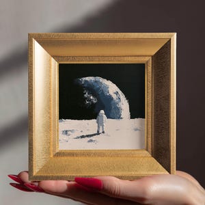 Puede incluir: Una pequeña pintura al óleo cuadrada en un marco dorado que representa a un astronauta de pie sobre una superficie lunar blanca. El fondo muestra un cielo negro y una gran forma de luna creciente blanca y texturizada con trazos gruesos.