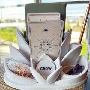 May include: A white lotus-shaped desk organizer with a speckled texture. It holds a green notebook, two tarot cards featuring a sun illustration, a small circular disc with the word GROW, copper-colored wire earrings, and a purple crystal pendulum.