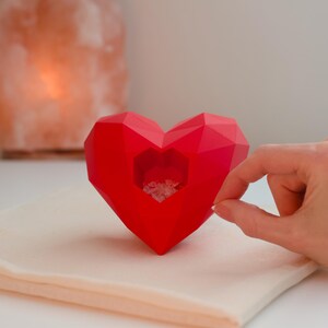 May include: A faceted red heart-shaped object with a smaller heart-shaped opening in the center, filled with crystals. The object is placed on a beige cloth. A hand is reaching towards the object.