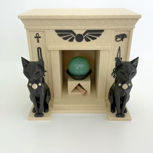 May include: A beige Egyptian-themed display with black cat statues on either side. The display features a central niche with a green sphere and a small, brown object. Decorative symbols and a winged sun disc are visible.