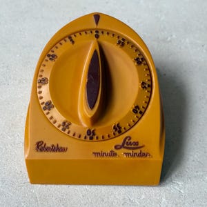 May include: Mustard-yellow vintage kitchen timer with a dark brown dial and a curved, oblong center. The timer has a circular dial with numbers and the words "Robertshaw" and "Lux minute minder" printed on the front.