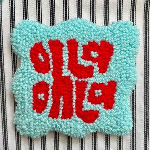 May include: A handmade, light blue patch with red text and a scalloped edge. The patch is made of textured material and is set against a striped background. The text is in a foreign language.
