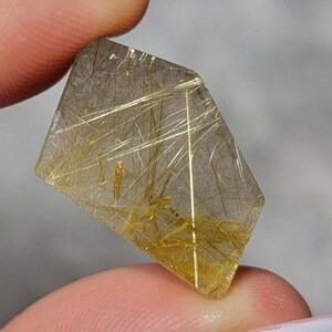 Puede incluir: Una piedra preciosa de cuarzo rutilado facetada de forma pentagonal, con un cuerpo claro y transparente lleno de numerosas inclusiones doradas finas, similares a agujas, que se entrecruzan en el interior.