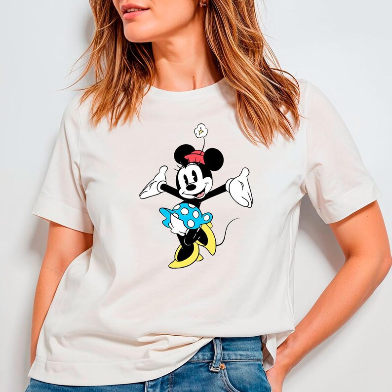May include: Cream-colored t-shirt featuring a cartoon graphic of Minnie Mouse. Minnie is depicted with a red hat, black ears, and a blue and white polka-dot skirt. The shirt has short sleeves and a crew neck.