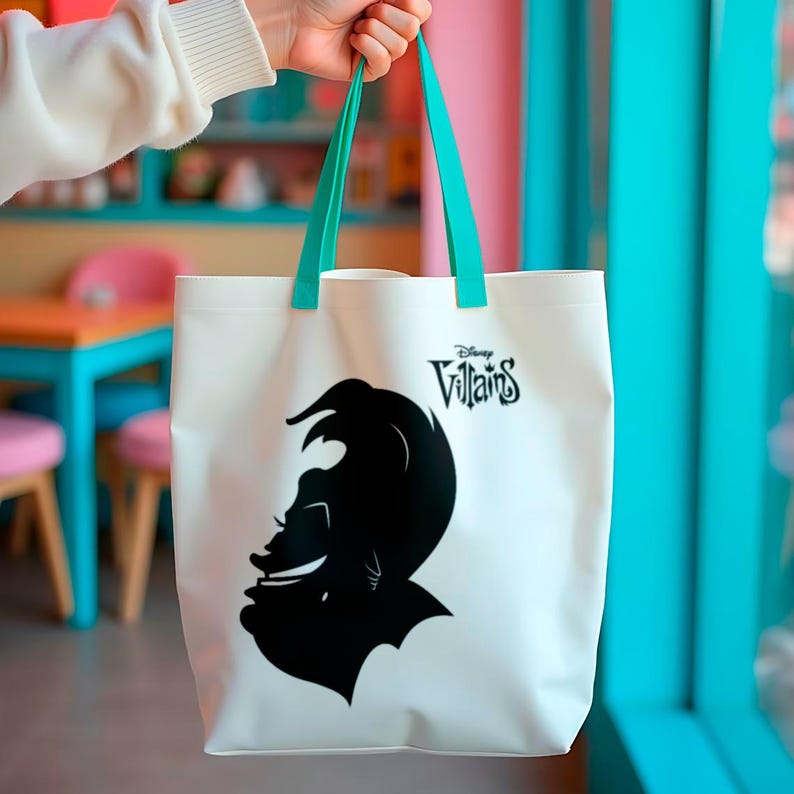 May include: White tote bag featuring a black silhouette of a Disney villain and the text "Disney Villains." The bag has teal green handles. The bag is held up by a person.