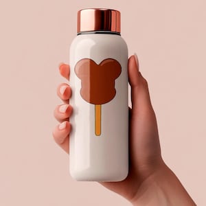 May include: White insulated water bottle with a copper-colored lid. The bottle features a brown cartoon popsicle design on the front. The bottle is held by a person's hand against a light pink background.