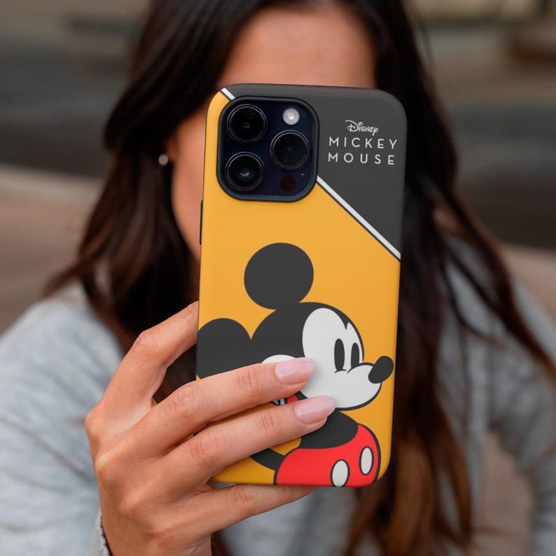 May include: Yellow and black phone case featuring Mickey Mouse. The case has a black top section with the Disney Mickey Mouse logo. The design includes a large Mickey Mouse graphic with red shorts. The phone case is held by a person.
