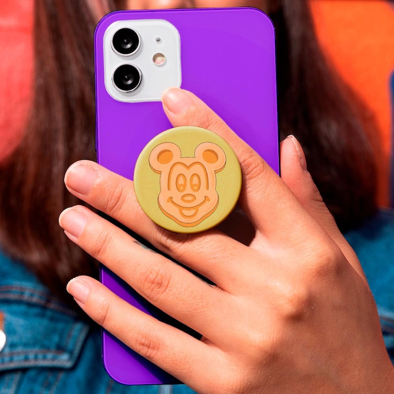 May include: A purple smartphone with a yellow pop socket featuring a smiling Mickey Mouse design. The pop socket is attached to the back of the phone, providing a secure grip.
