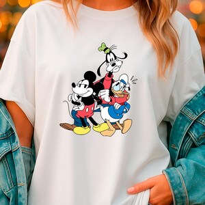 May include: White t-shirt featuring a colorful graphic of Mickey Mouse, Goofy, and Donald Duck. Mickey Mouse is in red shorts, Goofy in a red jacket, and Donald Duck in a blue sailor suit. The cartoon characters are hugging each other.