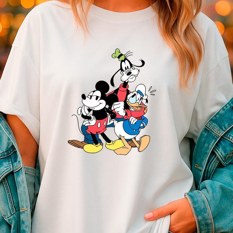 May include: White t-shirt featuring a colorful graphic of Mickey Mouse, Goofy, and Donald Duck. Mickey Mouse is in red shorts, Goofy in a red jacket, and Donald Duck in a blue sailor suit. The cartoon characters are hugging each other.