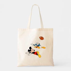 May include: Off-white tote bag featuring a cartoon illustration of Mickey Mouse and Donald Duck playing football. Mickey is in a red shirt, black shorts, and yellow shoes. Donald is in a blue shirt and yellow pants. A brown football is in the air.
