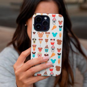 May include: White phone case featuring a repeating pattern of cartoon food and character icons. The design includes ice cream cones, pizza slices, and Mickey Mouse-shaped treats in various colors. The case is held by a person.