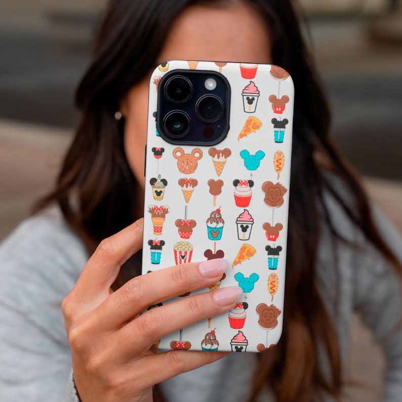 May include: White phone case featuring a repeating pattern of cartoon food and character icons. The design includes ice cream cones, pizza slices, and Mickey Mouse-shaped treats in various colors. The case is held by a person.